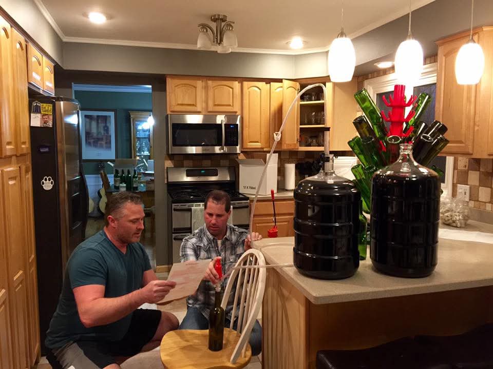 Russ (left, in a teal t-shirt) and Todd (right, in a plaid shirt) sitting at the kitchen island bottling their first homemade wine, with a siphon running into a bottle, two full carboys, and a bottle tree of green wine bottles drying behind them